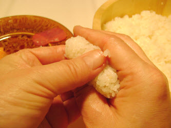 Take thumb and middle finger of right hand on either side of oval rice log and form a more rounded rectangle shape; at the same time press the thumb of left hand into rice log to form a depression